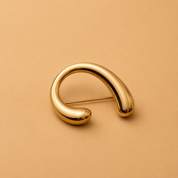A polished gold crescent-shaped brooch with a pin mechanism, displayed on a warm beige background.