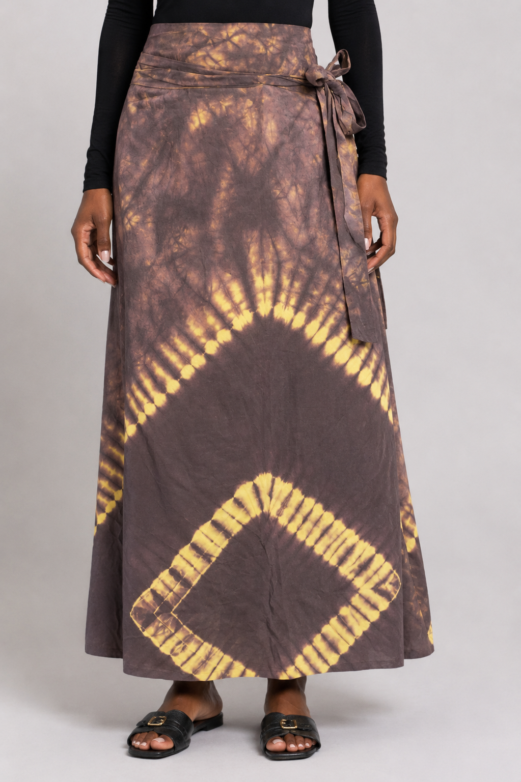Woman wearing a brown and yellow tie-dye wrap maxi skirt with a side tie waist and black leather slide sandals.